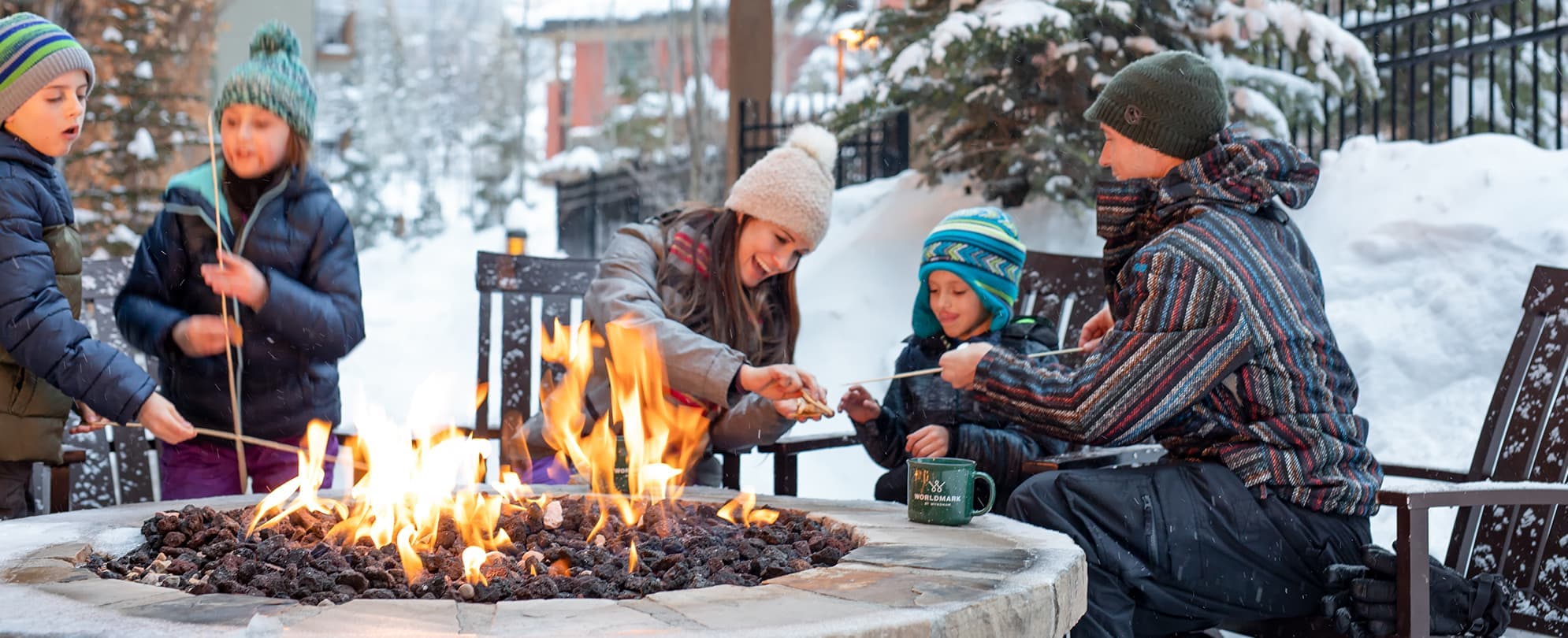 Family of five bundled up from the snow toasting marshmallows at an outdoor fire pit
